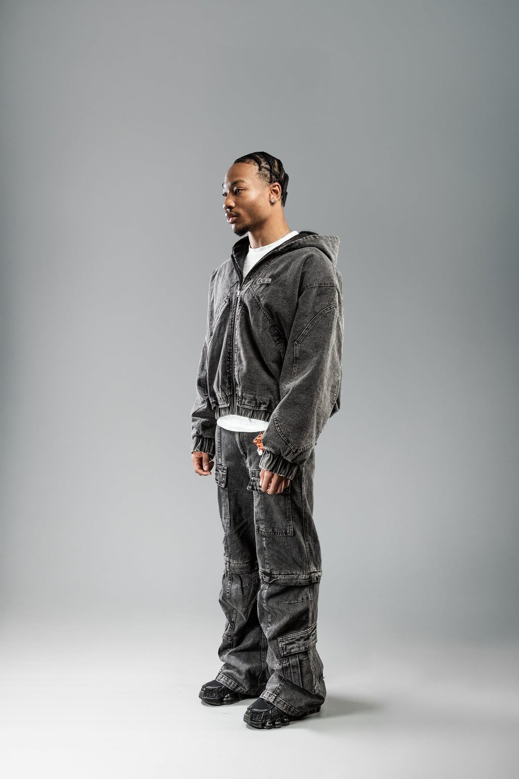 Die Hose wird mit einem offenen Hoodie getragen, das linke Bein leicht vorgedreht. Sichtbar sind die großen Taschen mit Klappen sowie der leicht aufgeraute Denim-Stoff in dunkler Waschung.