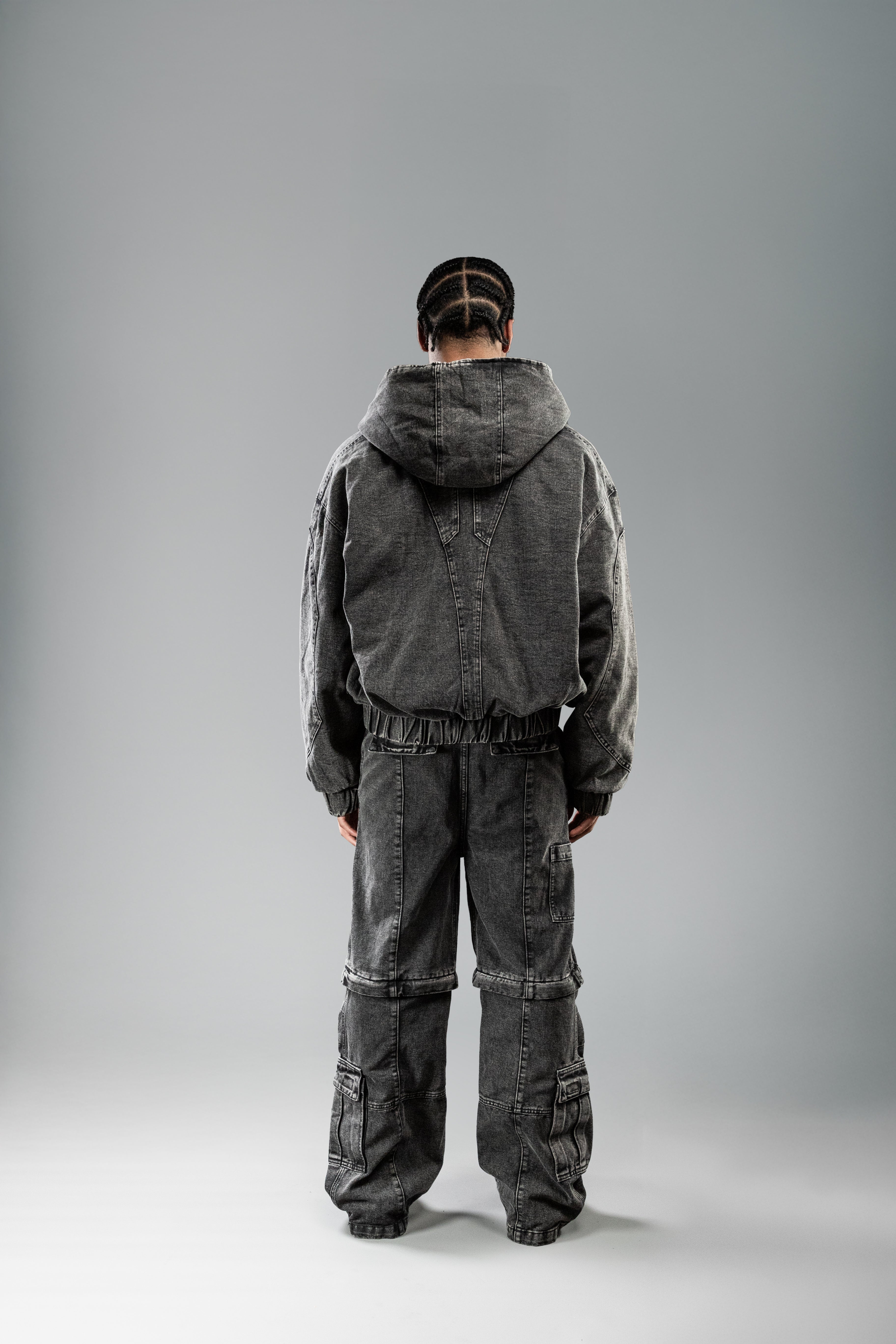 Rückansicht einer Person in schwarzer Kapuzenjacke und passender Hose. Die Kapuze ist hochgezogen, der Rücken zeigt eine markante Teilungsnaht in V-Form.