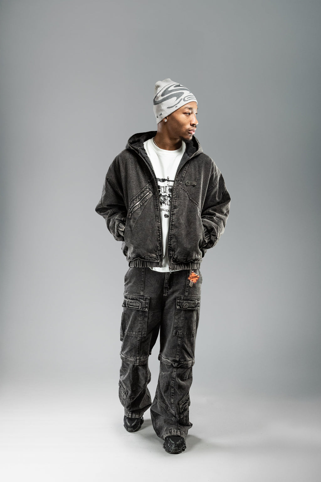 Person in schwarzer Denimjacke mit offener Kapuze, weißem Printshirt und passender Hose. Die Jacke ist vorne leicht geöffnet, das grafische Shirt darunter sichtbar.