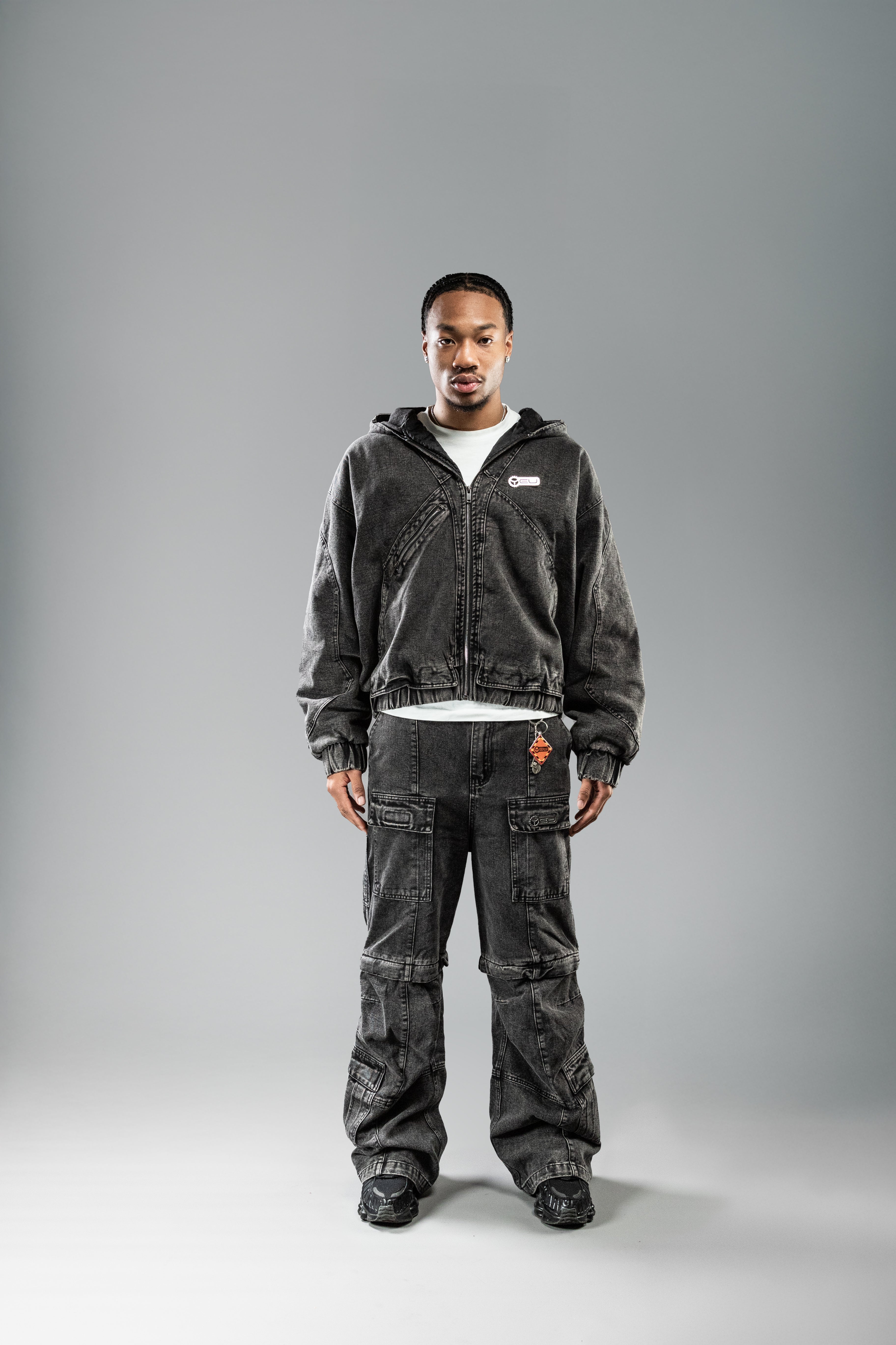 Die Hose wird mit einem offenen Zip-Hoodie getragen. Die Cargo-Elemente sind deutlich sichtbar, darunter zwei aufgesetzte Taschen am Oberschenkel mit Labelpatch sowie Seitentaschen auf Hüfthöhe.