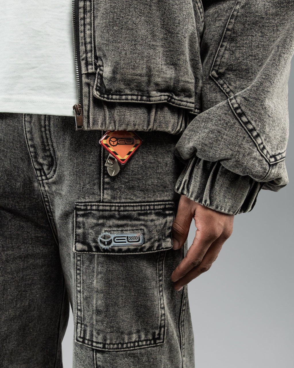 Nahaufnahme der rechten Beintasche der hellgrauen Hose mit aufgenähtem Gummipatch und einem auffälligen orangefarbenen Schlüsselanhänger mit Logo, der aus der Jackentasche hängt.
