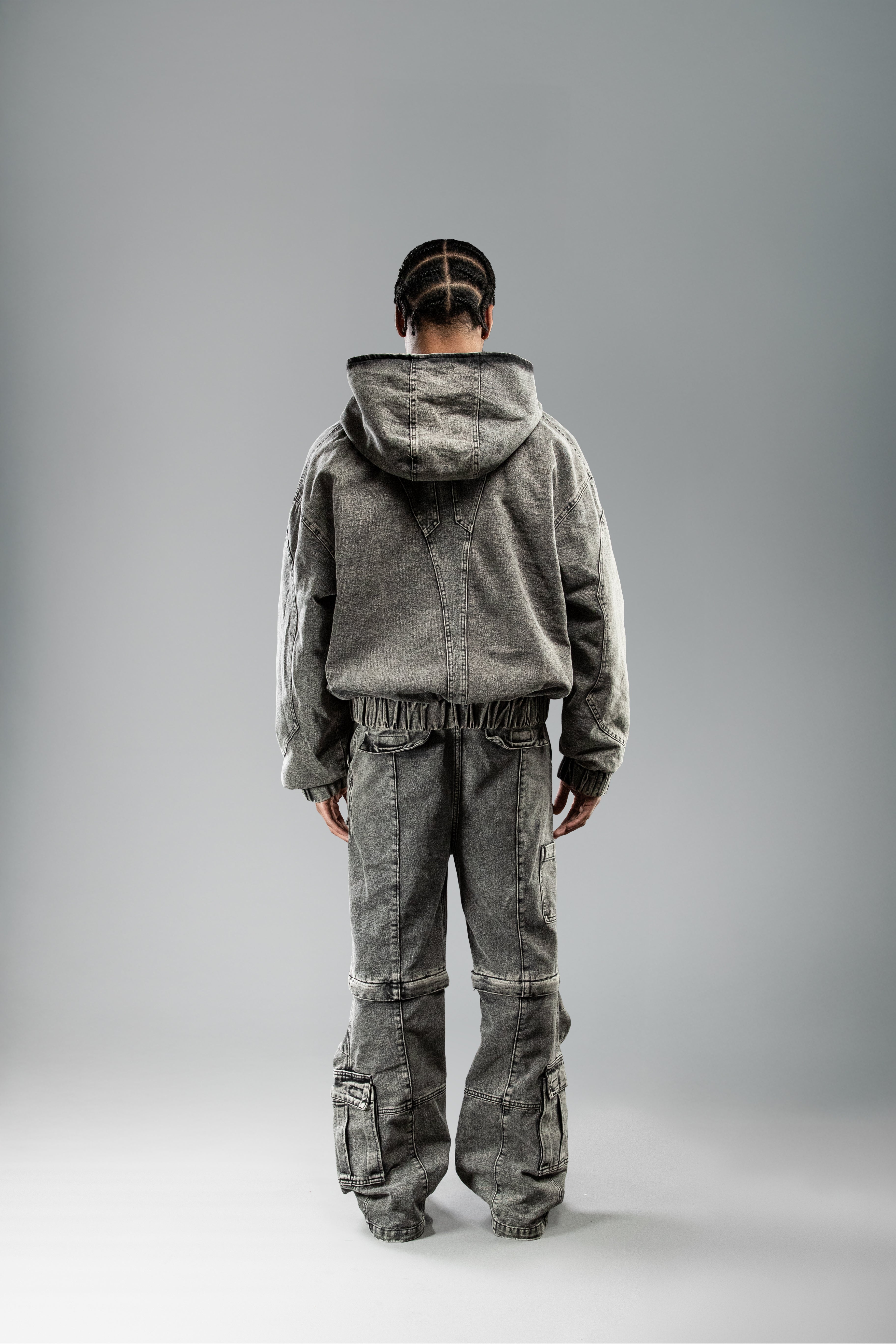 Rückansicht der hellgrauen Workwear-Hose am Model getragen, zusammen mit passender Jacke. Die Hose zeigt großflächige Rückseitenpanels, Teilungsnähte auf Kniehöhe sowie aufgesetzte Taschen an Gesäß und Beinen.