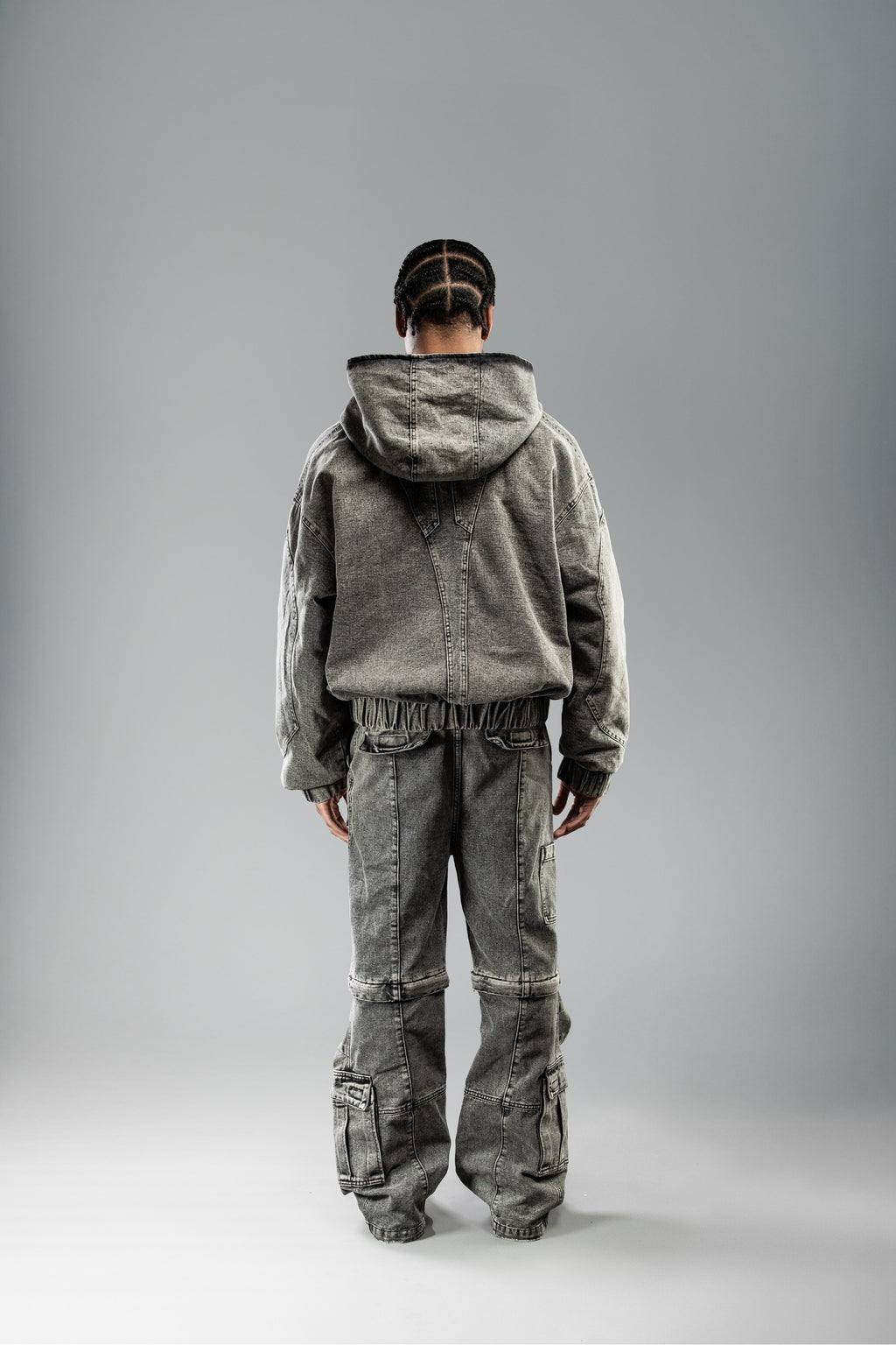 Rückansicht der Jacke und Hose an einem stehenden Model. Die Kapuze der hellgrauen Jeansjacke ist aufgesetzt, und im oberen Rückenbereich ist ein V-förmiges Teilungsnahtmuster zu sehen. Die Jacke ist kurz geschnitten mit elastischem Bund, passend zur Hose im gleichen Stil.