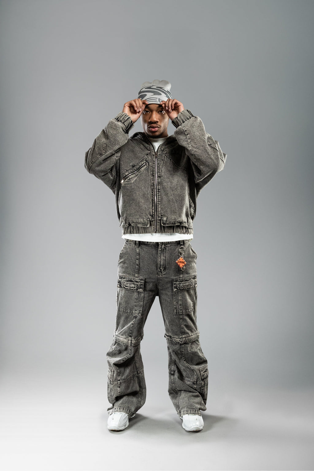 Person zieht sich eine beigefarbene Beanie mit grauem Wellenmuster und „YEU“-Schriftzug über den Kopf. Dazu wird ein graues Workwear-Set getragen.