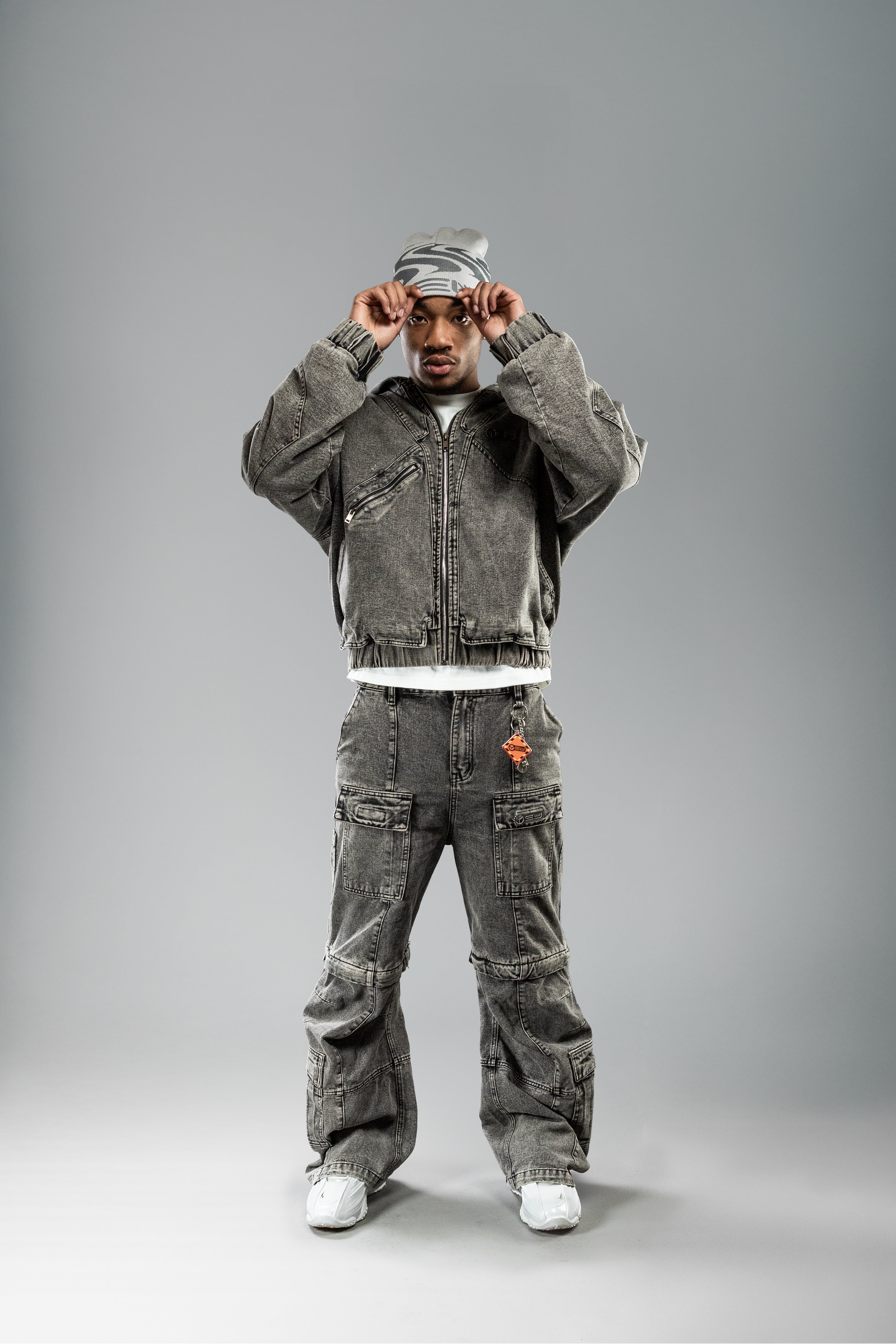 Person zieht sich eine beigefarbene Beanie mit grauem Wellenmuster und „YEU“-Schriftzug über den Kopf. Dazu wird ein graues Workwear-Set getragen.