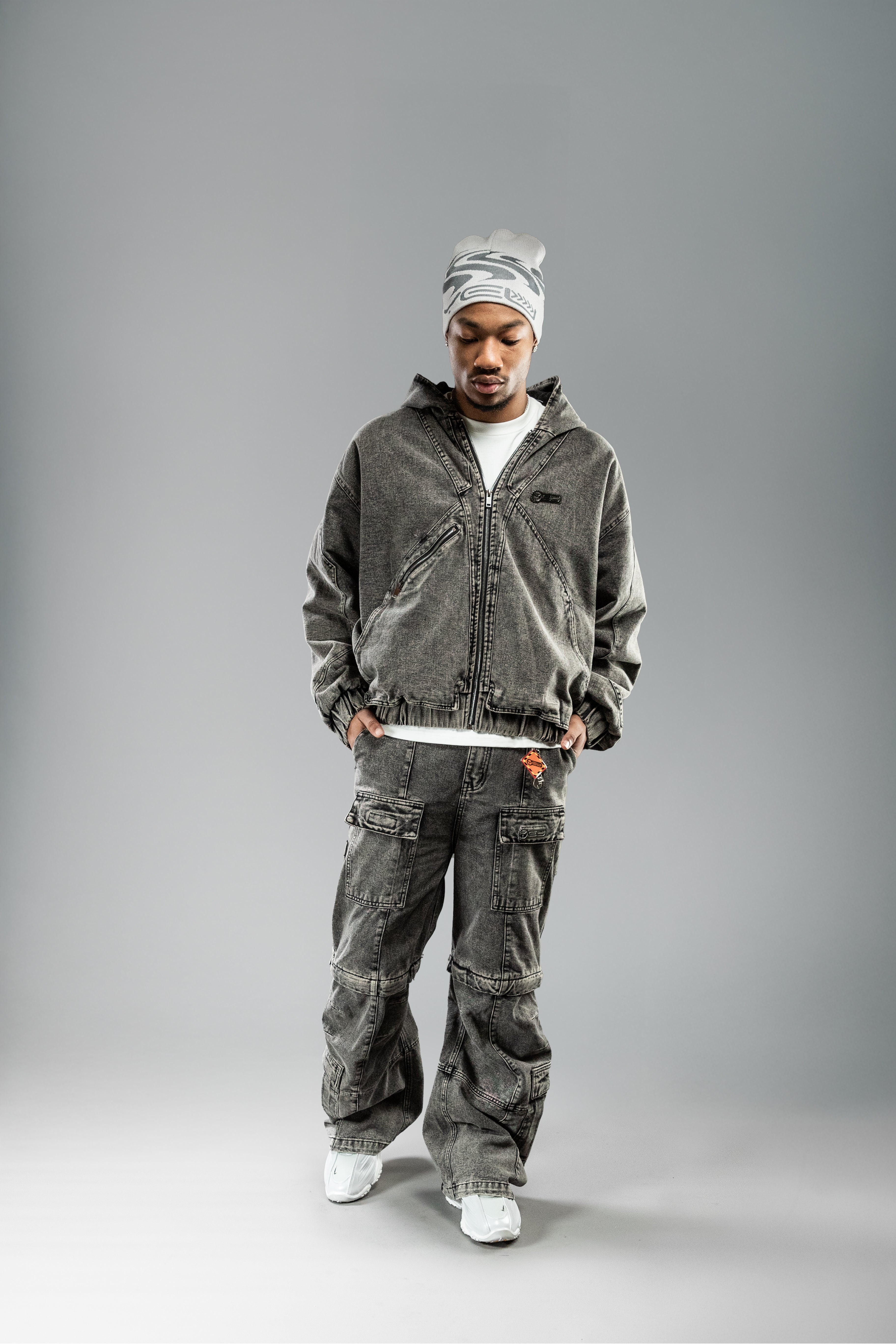 Person trägt eine beigefarbene Beanie mit grauem grafischem Wellenmotiv und „YEU“-Schriftzug. Dazu ein graues Workwear-Set mit Zip-Jacke.