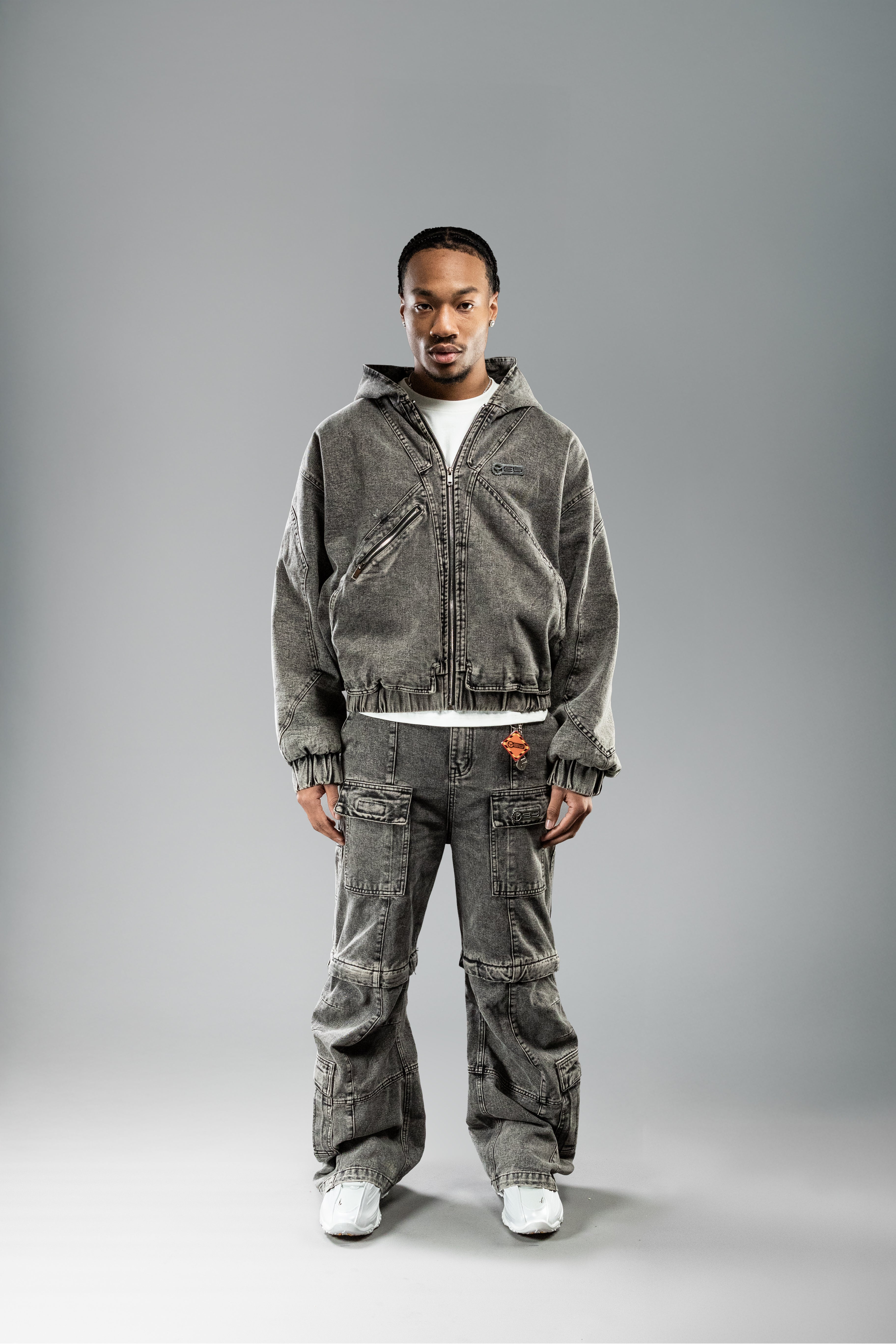 Frontalansicht eines Models in hellgrauem Jeans-Workwear-Set mit Kapuzenjacke. Die Jacke ist locker geschnitten, mit elastischen Bündchen und diagonal verlaufenden Nähten. Sichtbar sind der schräge Reißverschluss an der linken Brust, das CEU-Patch und ein weißes T-Shirt darunter. Die Hose hat breite Beine mit mehreren Taschen.