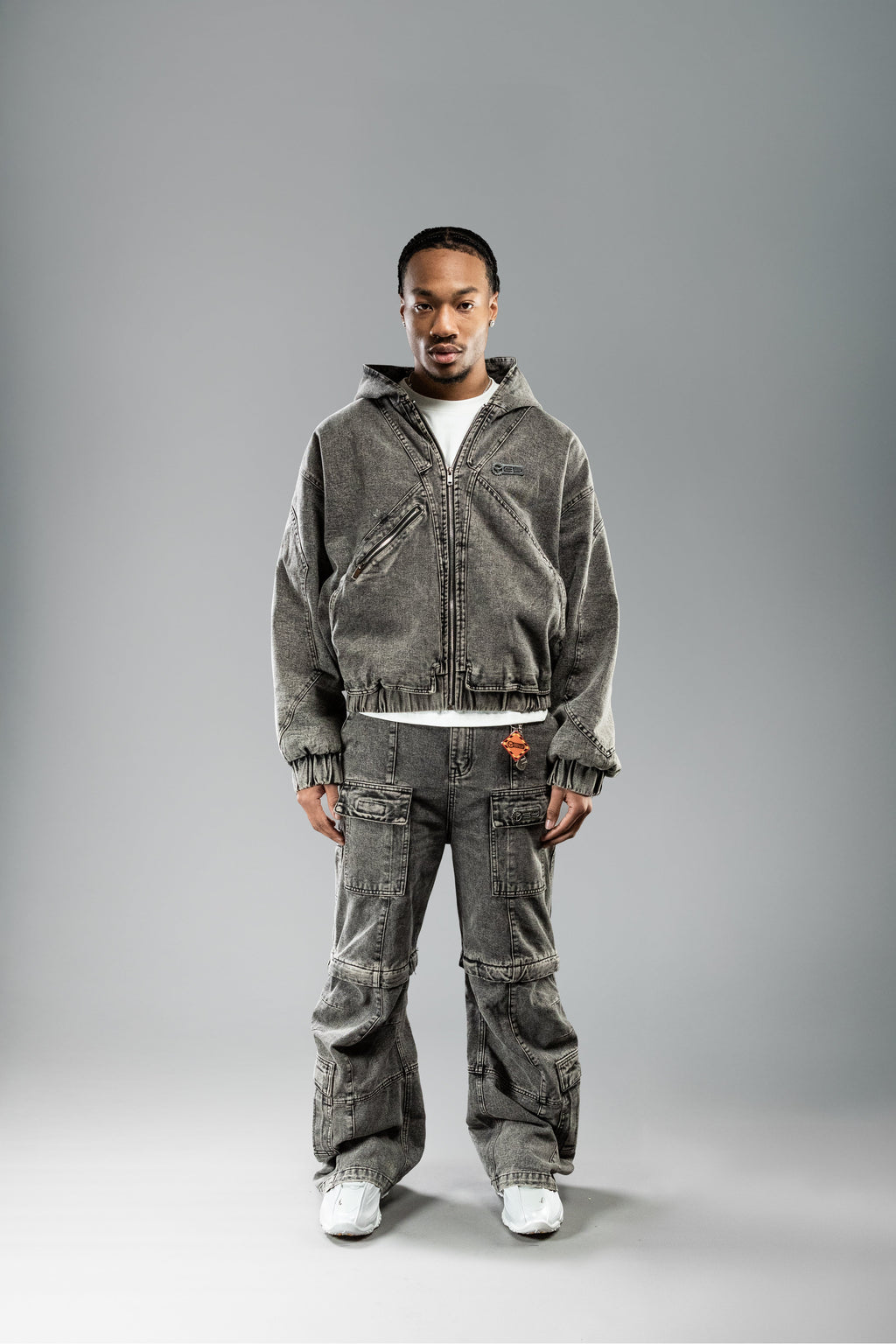 Frontalansicht eines Models in kompletter hellgrauer Workwear-Kombination mit Hose und Jacke. Die Hose hat eine weit geschnittene Form mit mehreren aufgesetzten Taschen und auffälliger Waschung.