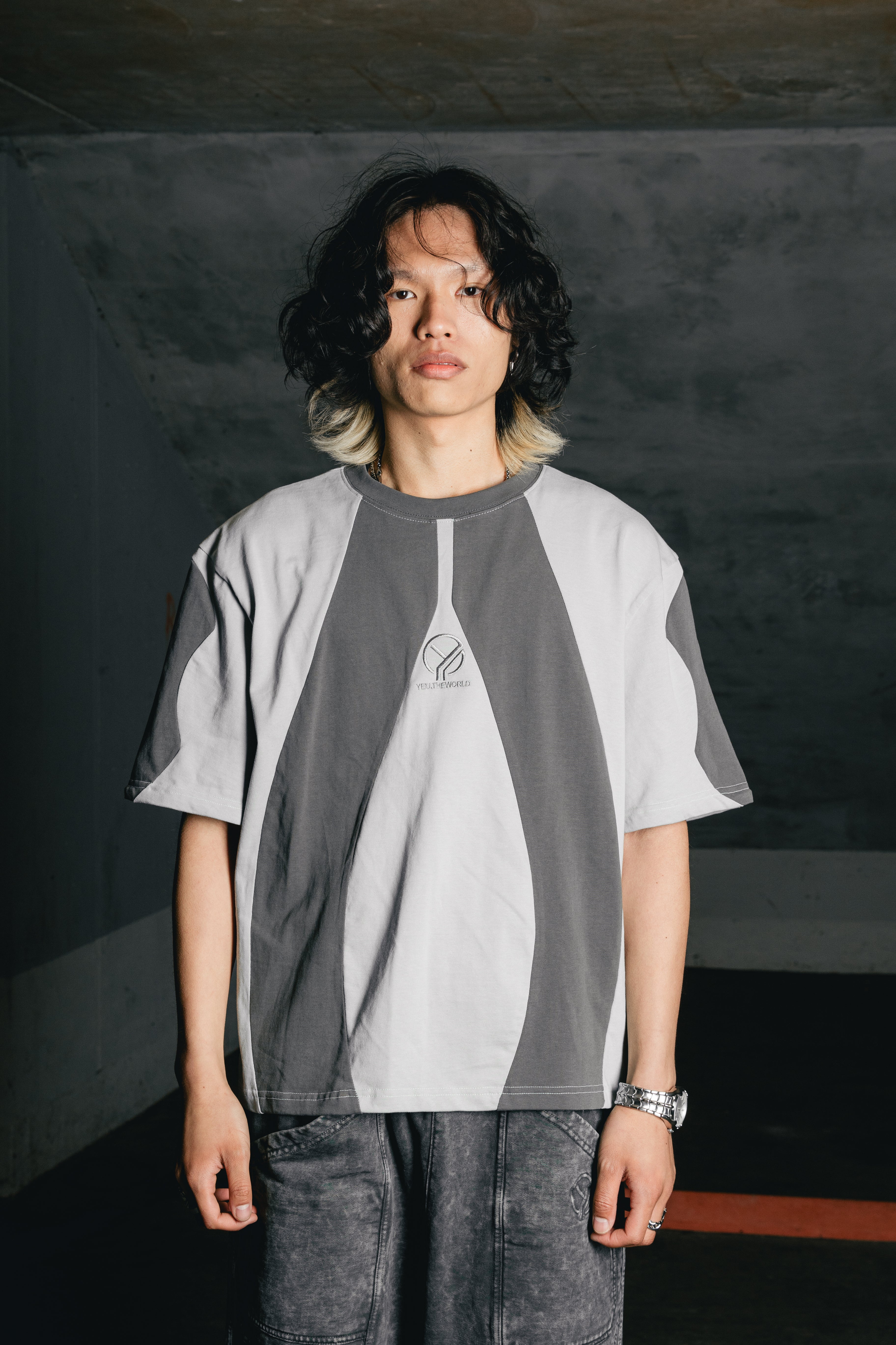 Person trägt ein zweifarbiges T-Shirt mit grafisch geschwungenen Farbflächen in Schwarz auf graugrünem Grund und zentraler Logo-Stickerei „YEU.THEWORLD“ auf der Brust.