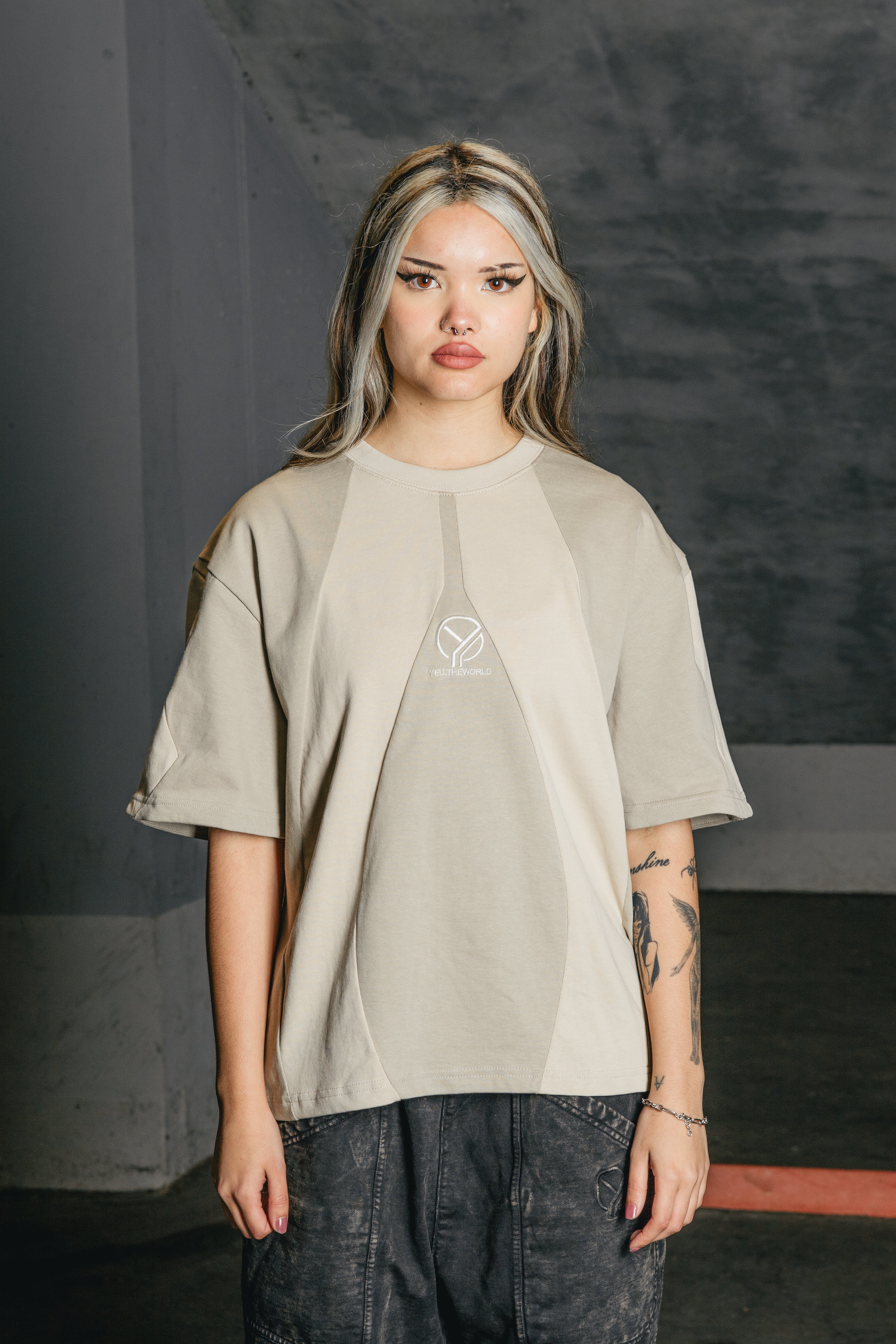 Tragefoto einer Person in dem cremefarbenen T-Shirt mit hellbeiger V-Form auf der Vorderseite und zentraler „YEU.THEWORLD“-Stickerei. Das Shirt ist locker geschnitten und wird zu verwaschenen schwarzen Pants getragen.
