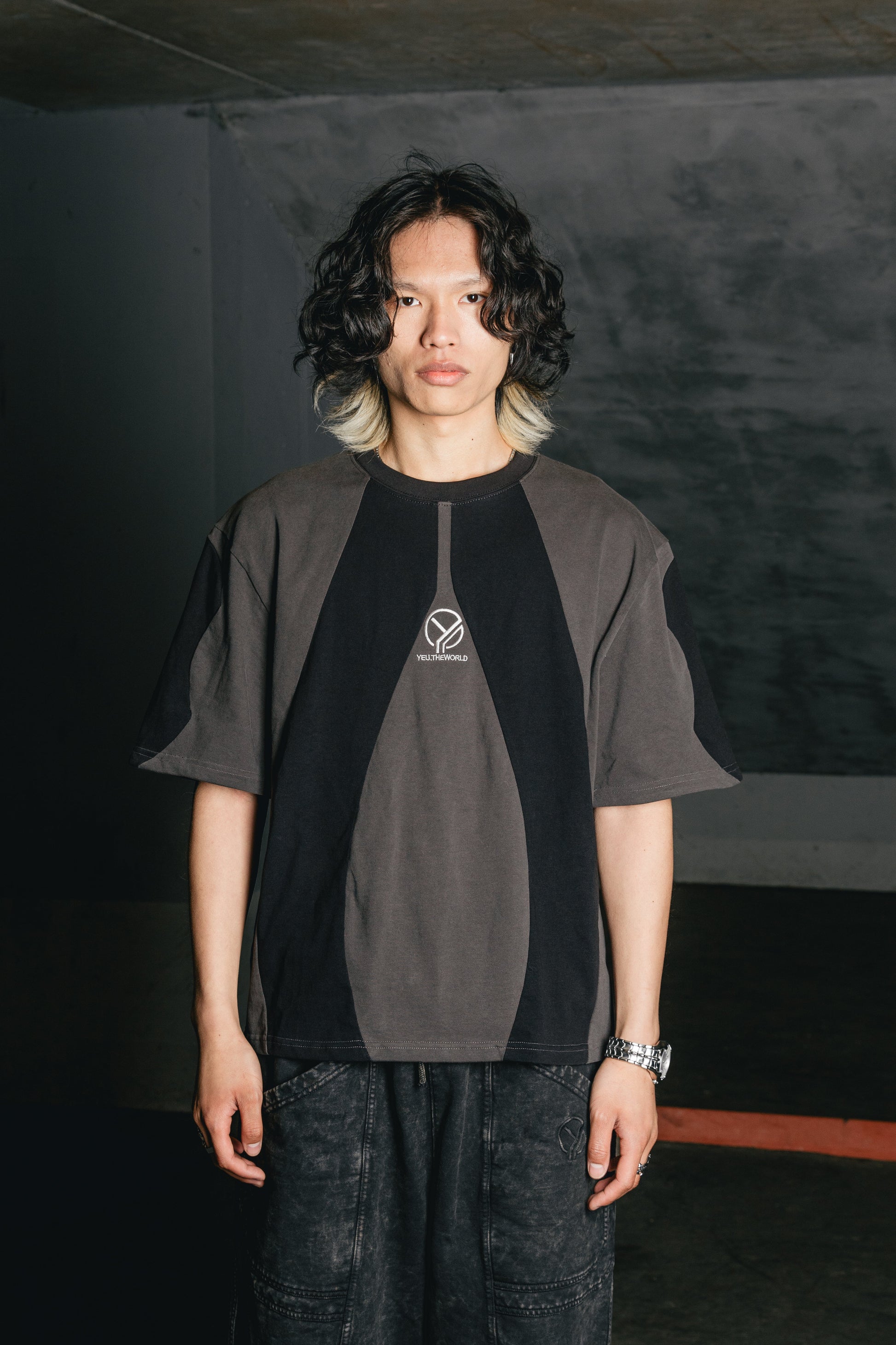 Das schwarze T-Shirt mit grauen, organisch geschwungenen Einsätzen wird von einem männlichen Model in einer neutralen Umgebung getragen. Die Stickerei „YEU.THEWORLD“ ist mittig auf der Brust platziert. Die geometrischen Panels betonen einen futuristischen Look.