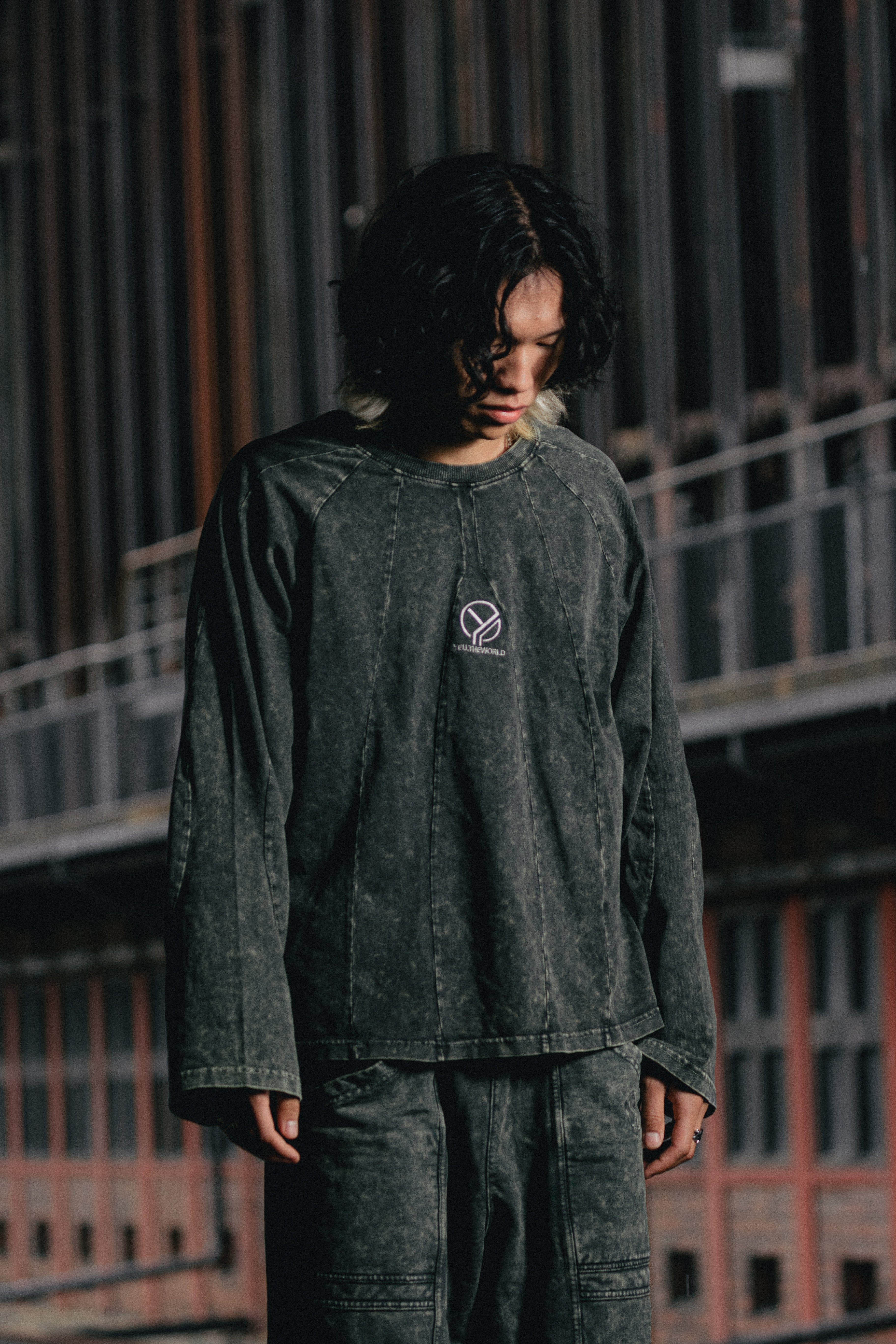 Tragebild in dunkler Urban-Umgebung: Eine Person trägt das langärmelige Shirt in verwaschenem Dunkelgrün mit vertikalen Ziernähten und mittiger Logo-Stickerei „YEU.THEWORLD“. Das Shirt wirkt locker geschnitten und wird mit passender verwaschener Hose kombiniert.